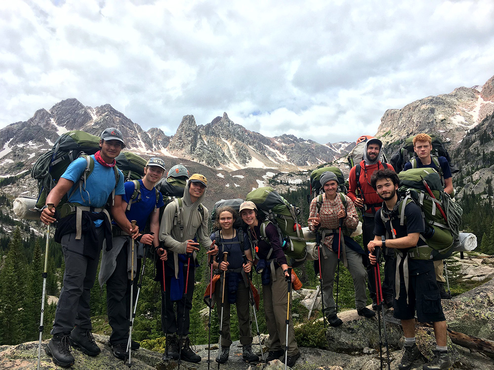 A group stands with backpacks on holding treking poles. A treed valley and mountain skyline are behind them.